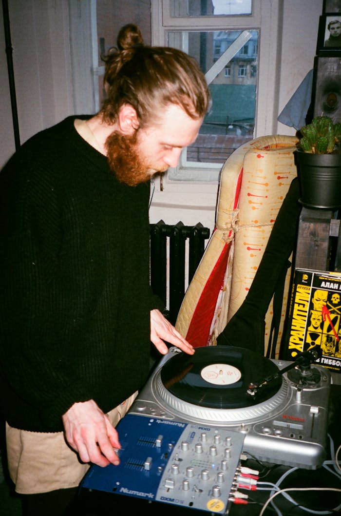 Man with beard and bun spinning vinyl on a turntable indoors in a cozy urban setting.