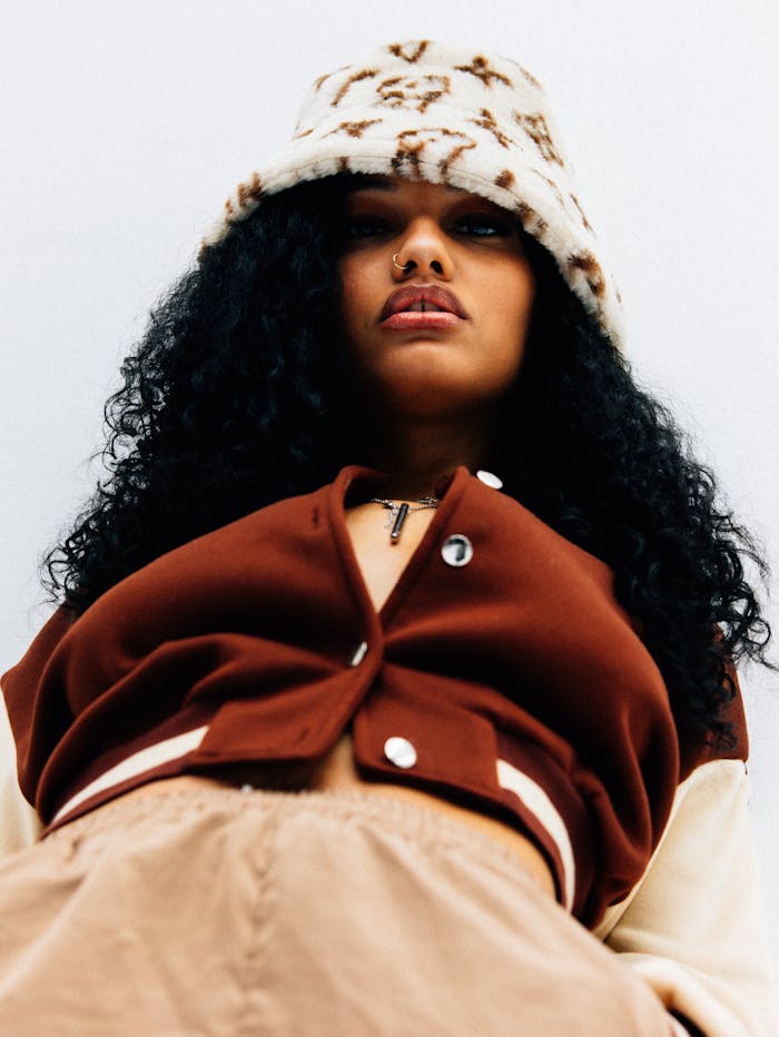 A fashionable woman wearing a brown outfit with a stylish hat and curly hair, looking confidently at the camera.